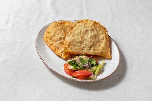 Chebureki stuffed with lamb, onion and suluguni cheese, lie on a plate. Nearby lies a vegetable salad, tomato, onion, garlic and cilantro. The food lies on an oval, light, ceramic plate. The plate stands on a light, linen fabric.