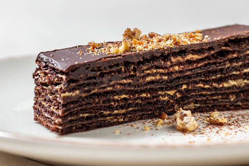 Chocolate puff cake with nuts. Wafer cakes soaked in cocoa with layers of nuts. Crushed walnuts are sprinkled on top of the cake. The cake lies on a light, round, ceramic plate. Nearby lies a scattered crushed walnut. The plate stands on a light, linen tablecloth.