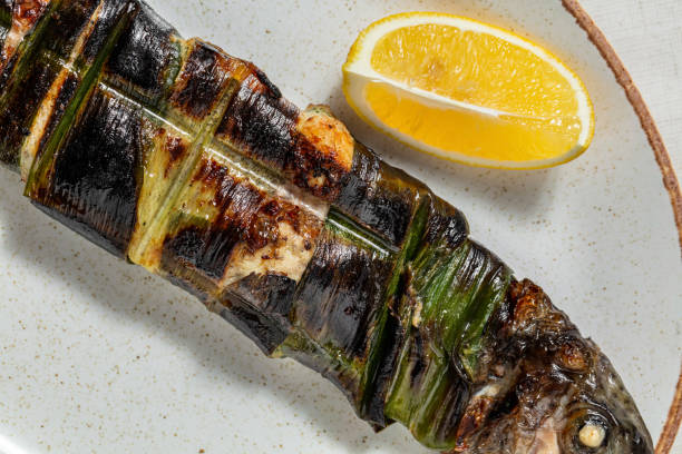 Grilled golden river trout baked in grape leaves with lemon, Svan salt and herbs. The trout lies on an oval, light, ceramic plate. Nearby lies a slice of lemon and a bunch of dill. The plate stands on a light, linen tablecloth. trucha dorada de río a la parrilla al horno en hojas de parra con limón, sal de svan y hierbas. la trucha se encuentra en una placa ovalada, ligera y de cerámica. cerca se encuentra una rodaja de limón y un manojo de eneldo. el plato se encuentra sobre