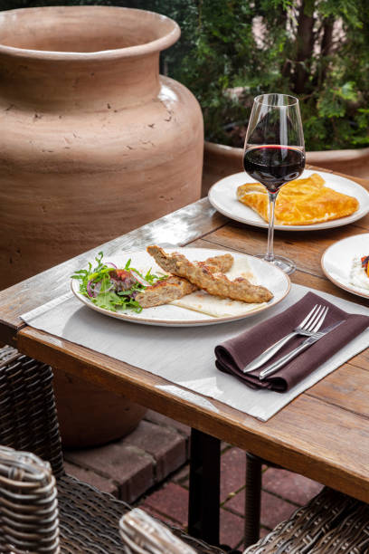 Chicken kebe lies in a light ceramic plate, next to it lies a salad. The plate is on a wooden table, next to it is a glass of red wine, then there is a plate of khachapuri. An armchair and plants can be seen next to the table. el kebe de pollo se encuentra en un plato de cerámica ligera, junto a él se encuentra una ensalada. el plato está sobre una mesa de madera, al lado hay una copa de vino tinto, luego hay un plato de khachapuri. un sillón y plantas se pueden ver al lado