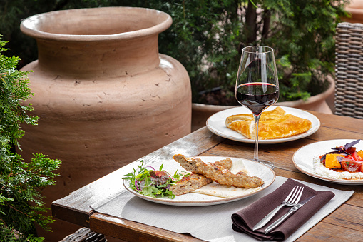 Chicken kebe lies in a light ceramic plate, next to it lies a salad. The plate is on a wooden table, next to it is a glass of red wine, then there is a plate of khachapuri. An armchair and plants can be seen next to the table.