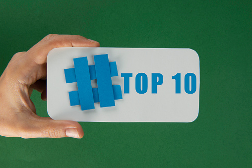 Hand is holding a blue hashtag symbol attached to a white smartphone like shape with top 10 text  in front of a green wall.