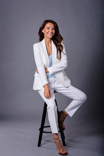 Full length studio portrait of an attractive woman relaxing at isolated dark background. Brunette haired mid aged female wearing white blazer and pants.