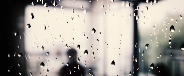 Rain drop on window glass of coffee shop and blurry city life background. Rainy season and blurry people city day life or bokeh night lights outside window. Coffee shop window covered with rain water