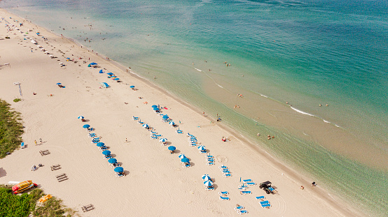 Vibrant Aerial Drone Views of Clear Teal Ocean Waves, People Relaxing Under Teal Blue Beach Umbrellas, Tall Hotels & Beach-Front Real Estate Buildings on Singer Island Beach, the Island East of Riviera Beach, Florida at Midday on Juneteeth 2023