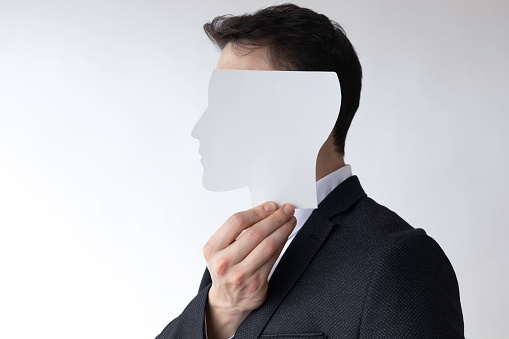 Unrecognizable businessman hiding his face using  white face paper mask in front of his face and is standing in front of white wall.