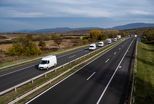 A large number of minivans move along the highway carrying goods for all people in the world. White modern delivery small shipment cargo courier van moving fast on motorway road to city urban suburb.