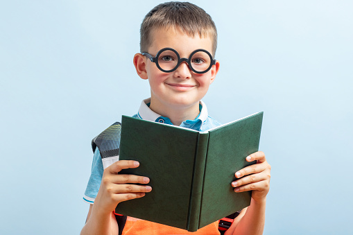 Positive elementary school nerd boy in eyeglasses holding open textbook and looking at camera against blue background
