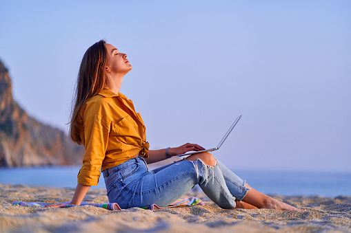 Satisfied millennial free freelancer woman using computer and sitting on sand beach by the sea. Enjoyment of dream office remote work concept