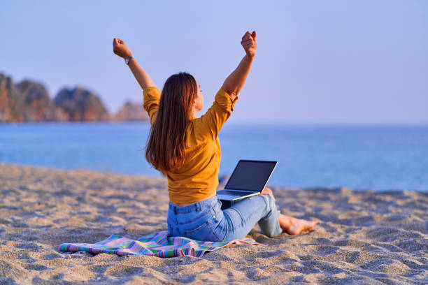 istock Satisfied millennial free freelancer woman with open arms using computer and sitting on sand beach by the sea. Enjoyment of dream office remote work concept 1500415632