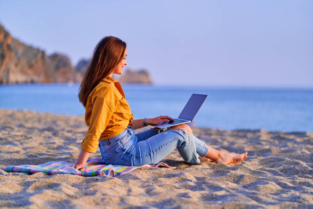 istock Young happy joyful smiling carefree satisfied millennial freelancer woman using computer and sitting on sand beach by the sea. Dream office remote work concept 1500415633