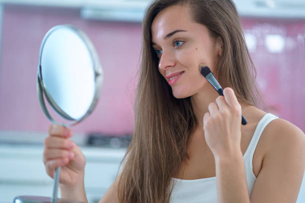 istock Beautiful attractive happy smiling brunette woman doing home makeup using a small round mirror and applying foundation on face with makeup brush in the morning 1500419917