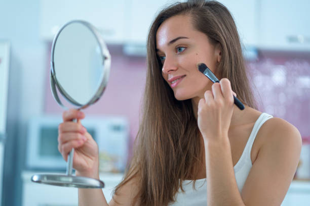 istock Beautiful attractive happy smiling brunette woman doing home makeup using a small round mirror and applying foundation on face with makeup brush in the morning 1500419925