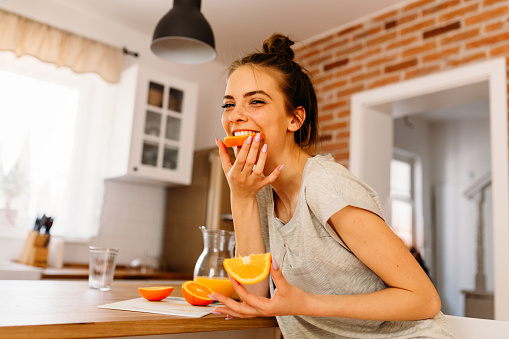 Cheerful young woman cutting slices of oranges and eating them for breakfast in the kitchen