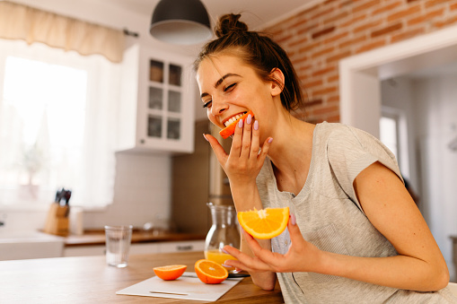 Cheerful young woman cutting slices of oranges and eating them for breakfast in the kitchen