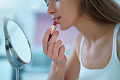 Beautiful attractive happy smiling brunette woman paints lips with red lipstick using a small round mirror during home makeup in the morning