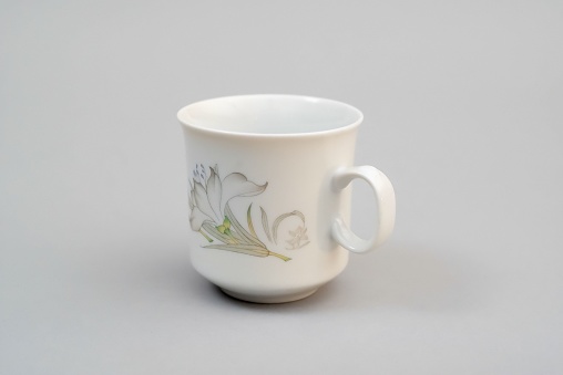 A white ceramic cup with a floral design, with a handle, standing on a light gray background