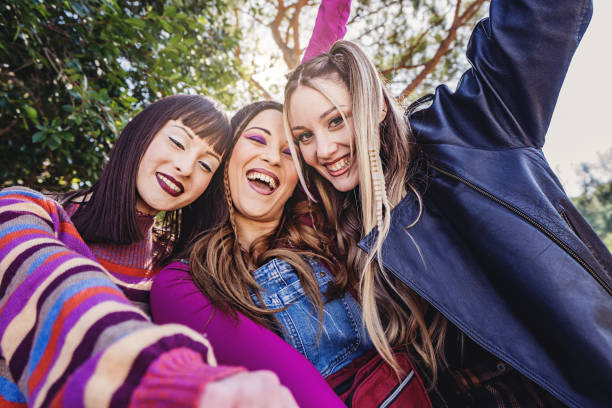Three Caucasian young women laughing, joking, singing, and posing for the camera in a public park. Bold makeup and casual 90s style clothing. Blonde and brunette friends, happy on a mild weather day.