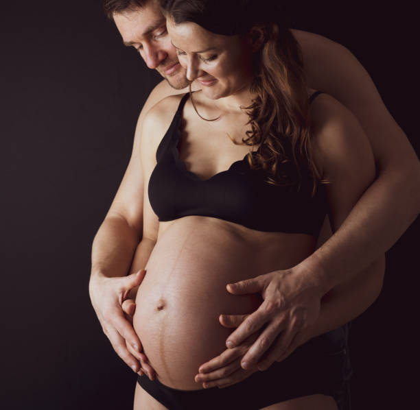 des parents fiers tenant le ventre rond nu d’une mère enceinte en sous-vêtements. dernier mois de grossesse - 36ème semaine. vue frontale. fond noir. - table chair white curve photos et images de collection