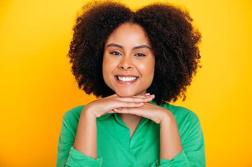 Close-up photo of an amazing charming african american or brazilian curly haired young woman with brown eyes, wearing a green shirt, looking at the camera, smiles friendly, isolated yellow background