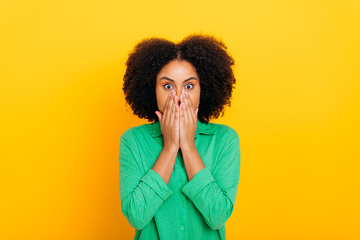 Scared worried brazilian or african american stylish curly haired woman, covering mouth with hands, looking at camera in shock, frightened, standing on isolated yellow background