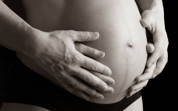 primer plano del vientre redondo desnudo de una madre embarazada en ropa interior sostenida por las manos de la madre y el padre. último mes de embarazo - semana 36. vista en ángulo lateral. fondo negro. sepia. - two parent family naked men couple fotografías e imágenes de stock