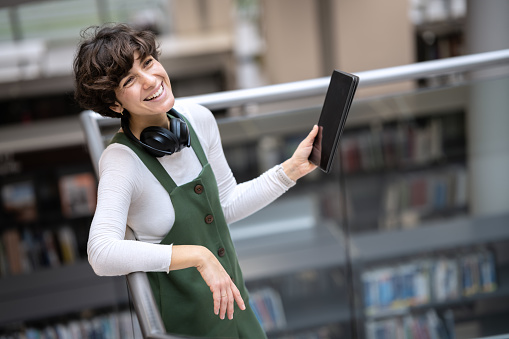 A young cute girl is standing on the top of the stairs with wifi headphones around her neck, holding a tablet, smiling and looking at someone, looking at the camera. Copy space.