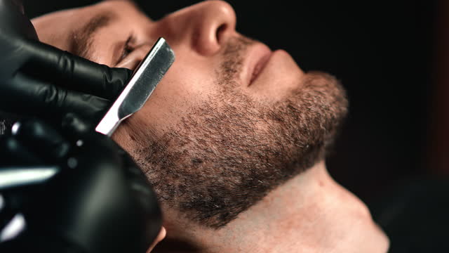 Male barber hands shaving customer beard use vintage sharp metal shaver isolated on black closeup