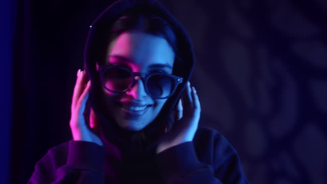 Portrait smiling hipster female listening music in headphones posing at dark neon backlit studio