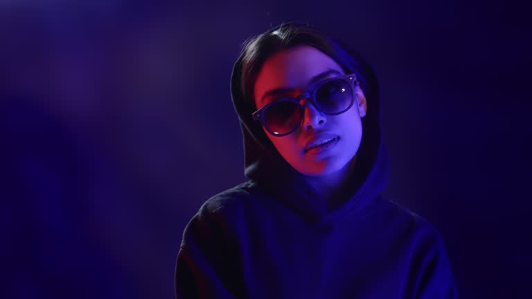 Fashion vogue hipster woman in hood sunglasses relaxing posing at dark backlit neon light studio