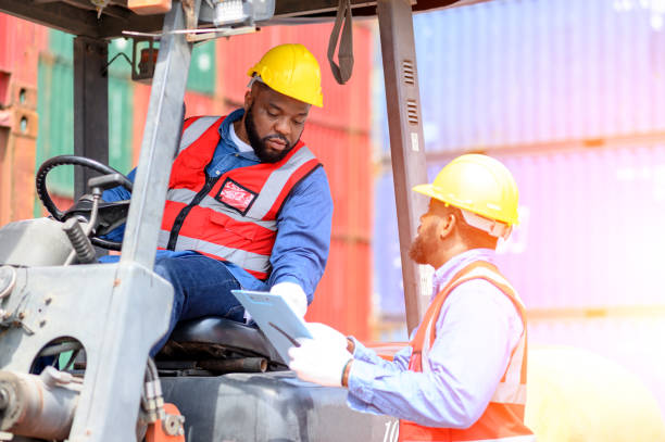 istock Two African American males in safety uniforms and helmets, container operating working with the forklift truck driver at commercial dock site. Cargo Ship Import Export Factory Logistic.. 1500473053
