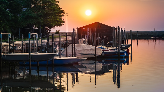 View of fishing pier at sunrise; boathouse, boats and pier refected in calm water