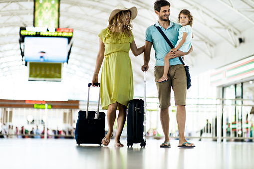 Young happy family pulling their luggage while walking down the airport. Mother is pregnant.