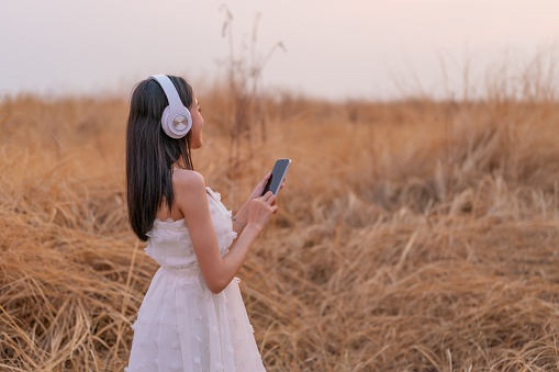 Photo of a young Asian woman in a white dress finds solace in nature's embrace. With headphones on, she listens to music, harmonizing with the serene sunset and the beauty of the surrounding field