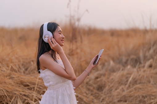 Photo of a young Asian woman in a white dress finds solace in nature's embrace. With headphones on, she listens to music, harmonizing with the serene sunset and the beauty of the surrounding field