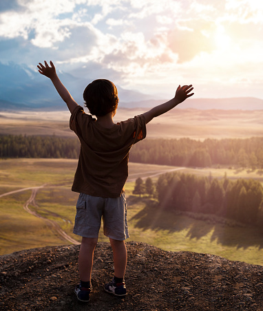 A little boy stands on top of a mountain with his hands up and looks at the sunset
