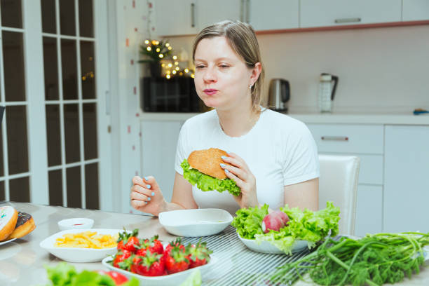 problèmes de boulimie, jeune femme mangeant un hamburger dans la cuisine avec une table pleine de nourriture différente - food measuring hamburger dieting photos et images de collection