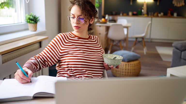 Young Caucasian woman, a university student, studying online and eating her healthy oatmeal breakfast.