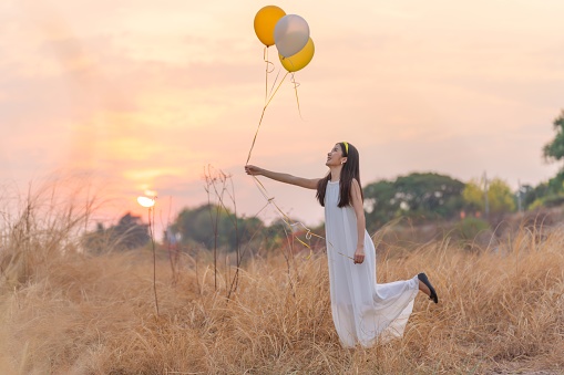 In a wide field of dry grasses during sunset, a young Asian lady in a white dress holds a cluster of colorful balloons, exuding pure joy and delight.
