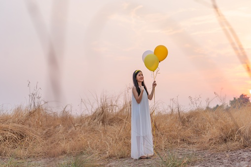 In a wide field of dry grasses during sunset, a young Asian lady in a white dress holds a cluster of colorful balloons, exuding pure joy and delight.