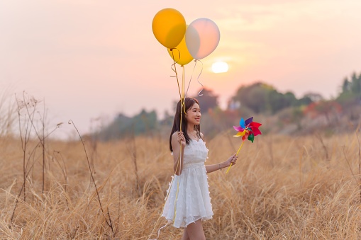 In a serene field of tall grasses, a young Asian lady in a white dress embraces a moment of whimsy. Holding windmill toys, she revels in the playful dance of colorful balloons, creating an enchanting scene of joy and lightheartedness