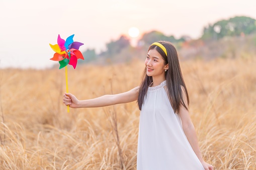 midst a wide field of dry grasses during sunset, a young Asian lady dons a white dress and holds a colorful windmill toy. Her delightful expressions and carefree wanderings epitomize the playful spirit and joy found within the enchanting beauty of the golden fields.