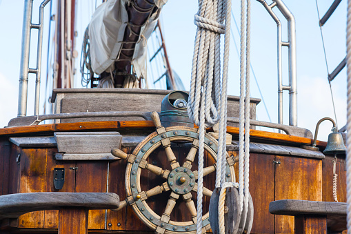 Old wooden sailing ship, closeup. Wooden steering wheel, ropes, rope ladder and bronze bell.