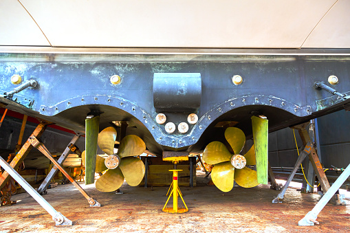 The back of a large motor yacht, standing on wooden blocks in a dry dock. Two propellers and zinc protective inserts.