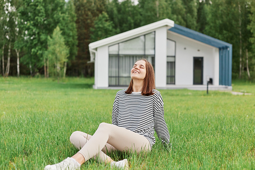 Relaxed enjoyable woman sitting on ground on grass against background of modern house resting in yard looking up with happy smile resting enjoying sunny summer day.