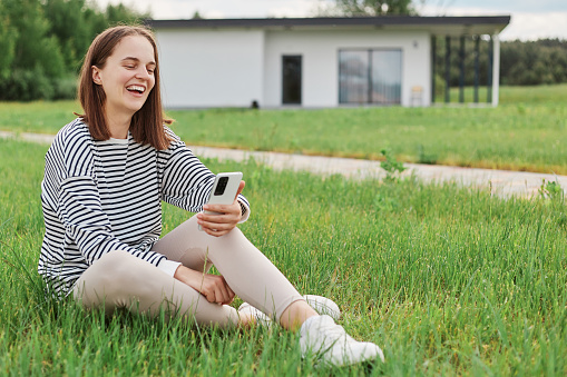 Beautiful attractive caucasian woman using cell phone sitting on grass on yard with big house on background having video call laughing talking with friends spending free time.