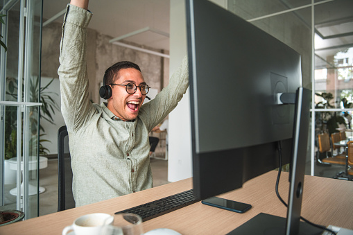 Witness a moment of celebratory success as a joyful Hispanic male, seated at her desk in a modern call center, raises his hands in triumph. With his desktop computer as his ally, he celebrates landing a valuable client, reflecting the dedication and skill she brings to his work in the dynamic call center environment.