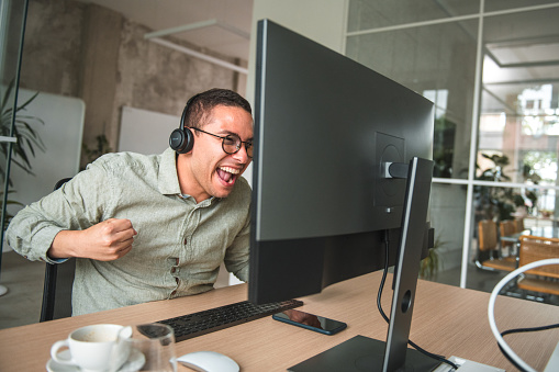 Experience the perfect blend of success and joy as a thrilled Hispanic male employee celebrates landing a client in a contemporary call center. Seated at his desk and utilizing his desktop computer, he expresses his excitement with hands raised high. His hard work, skills, and commitment have paid off, reflecting his exceptional performance in securing new clients within the modern office space.