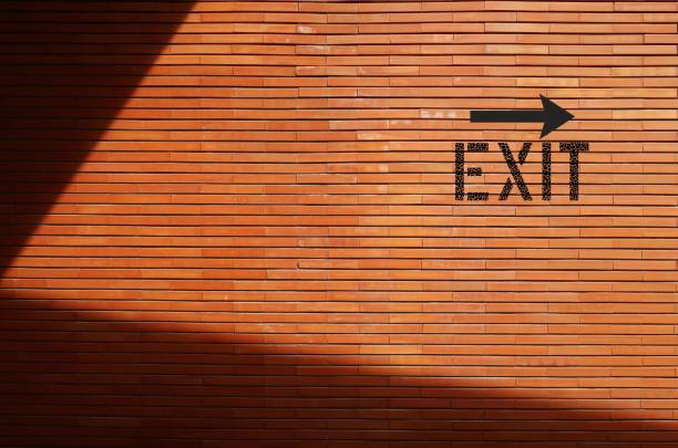 exit direction sign on copy space brick shadow wall- shows way out of public building - finding way out from trouble, departure from toxic relationship - escaping the rat race imagens e fotografias de stock