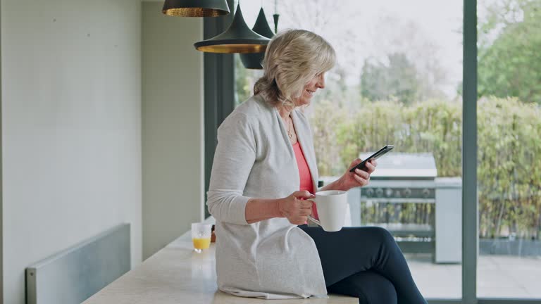 Senior woman reading phone news, mobile app and social media during morning coffee in home kitchen. Relax retirement with smartphone and connection in apartment